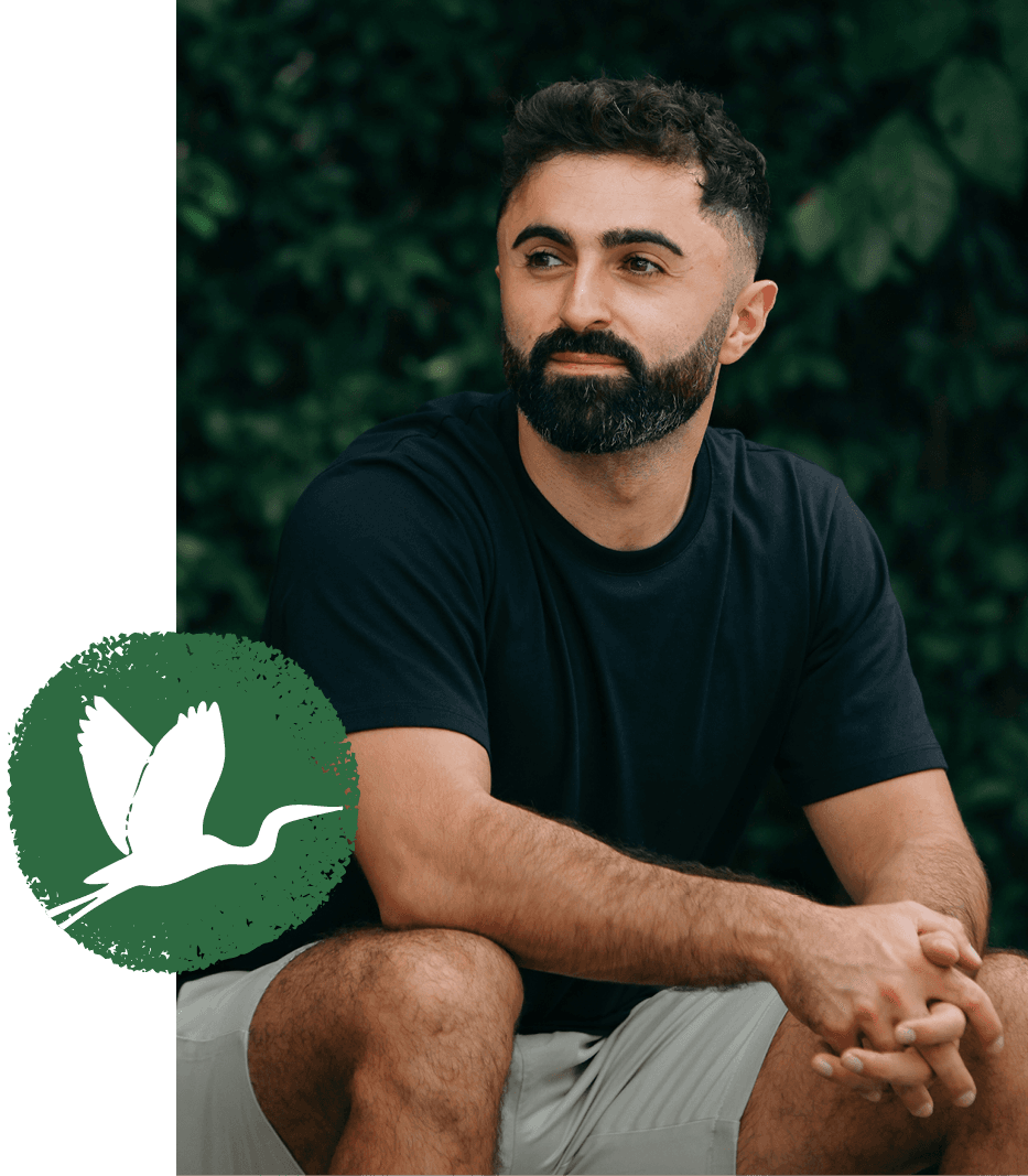 A man sitting outdoors in front of greenery, with the St. Christopher's heron emblem